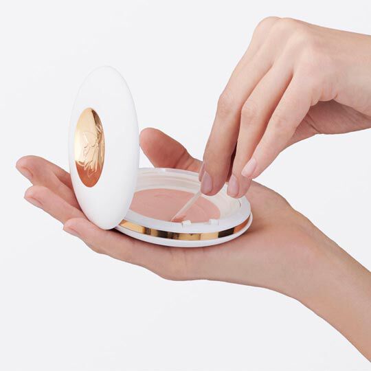Eine Hand halt einen runden, weißen Makeup-Spiegel mit goldener Verzierung, wahrend eine andere Hand einen Pinsel oder Applikator in die Dose eintaucht.