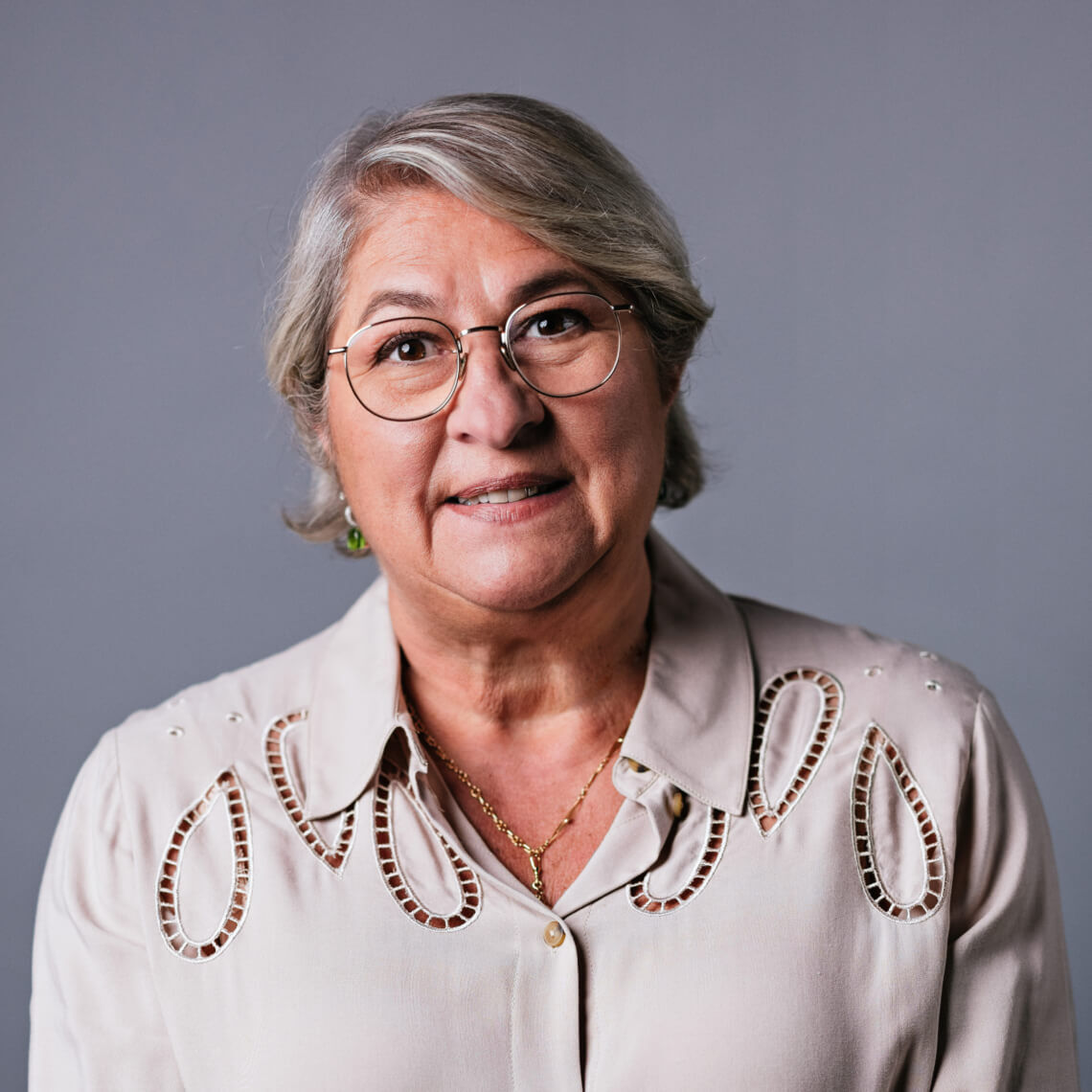 Altere Frau mit Brille und lachelndem Gesicht, tragt eine helle Bluse mit besonderen Details, vor grauem Hintergrund.