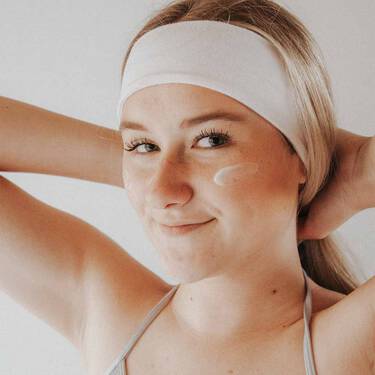 Dekoratives Bild: Eine junge Frau lachelt in die Kamera, hat ein Haarband auf und macht sich fur ihre Skincare Routine bereit