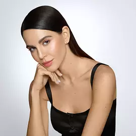 Eine Frau mit langen, glatten Haaren und sanftem Make-up posiert mit einer Hand an ihrem Kinn. Sie tragt ein schwarzes Top und blickt freundlich in die Kamera. Der Hintergrund ist hell und neutral.