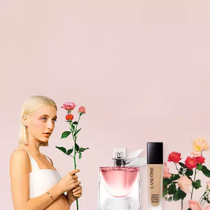 Eine blonde Frau in einem weißen Oberteil halt eine Rose und blickt direkt in die Kamera. Auf der rechten Seite sind verschiedene Lancome-Produkte und Blumen abgebildet, vor einem sanften rosa Hintergrund.