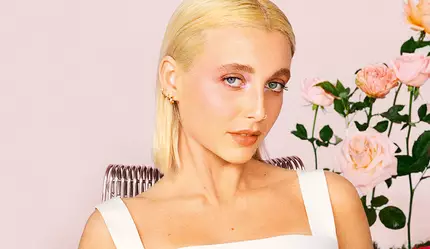 Eine Person mit blonden Haaren tragt ein weißes Oberteil und sitzt vor einem rosa Hintergrund mit Rosen.