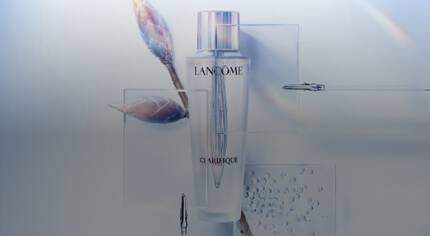 Flasche des Lancome Clarifique-Produkts vor einer stilisierten, hellen Hintergrundgestaltung mit zarten Zweigen und Wassertropfen.