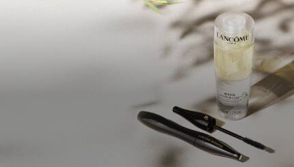 Alt-Text: Ein Makeup-Produkt von Lancome, bestehend aus einer Flasche Clean & Care und einem Mascara mit abnehmbaren Deckel, auf einem spiegelnden Untergrund. Im Hintergrund sind grune Pflanzenblatter sichtbar.