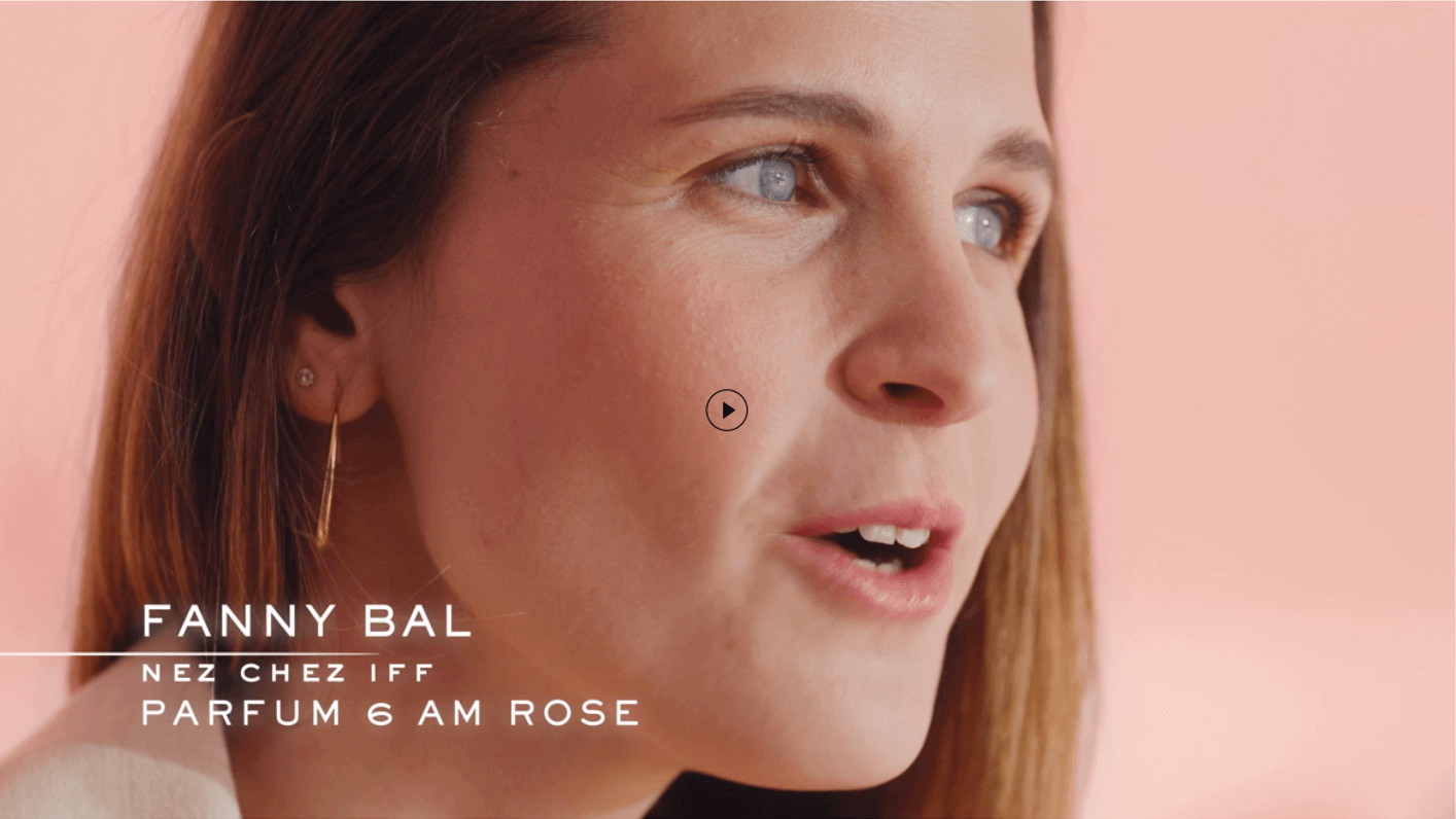 Porträt einer Frau mit langen Haaren, die über das Parfum '6 AM Rose' spricht, vor einem sanften pinken Hintergrund.