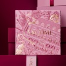 Rosa Lancôme-Geschenkbox mit festlichem Muster vor einem burgunderroten Hintergrund.