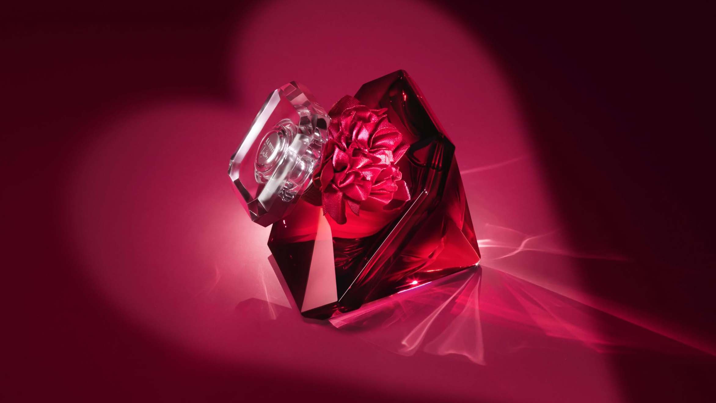 Ein eleganter, roter Parfümflakon in Form eines Diamanten, mit einer blütenförmigen Verschlusskappe, vor einem rosaroten Hintergrund.